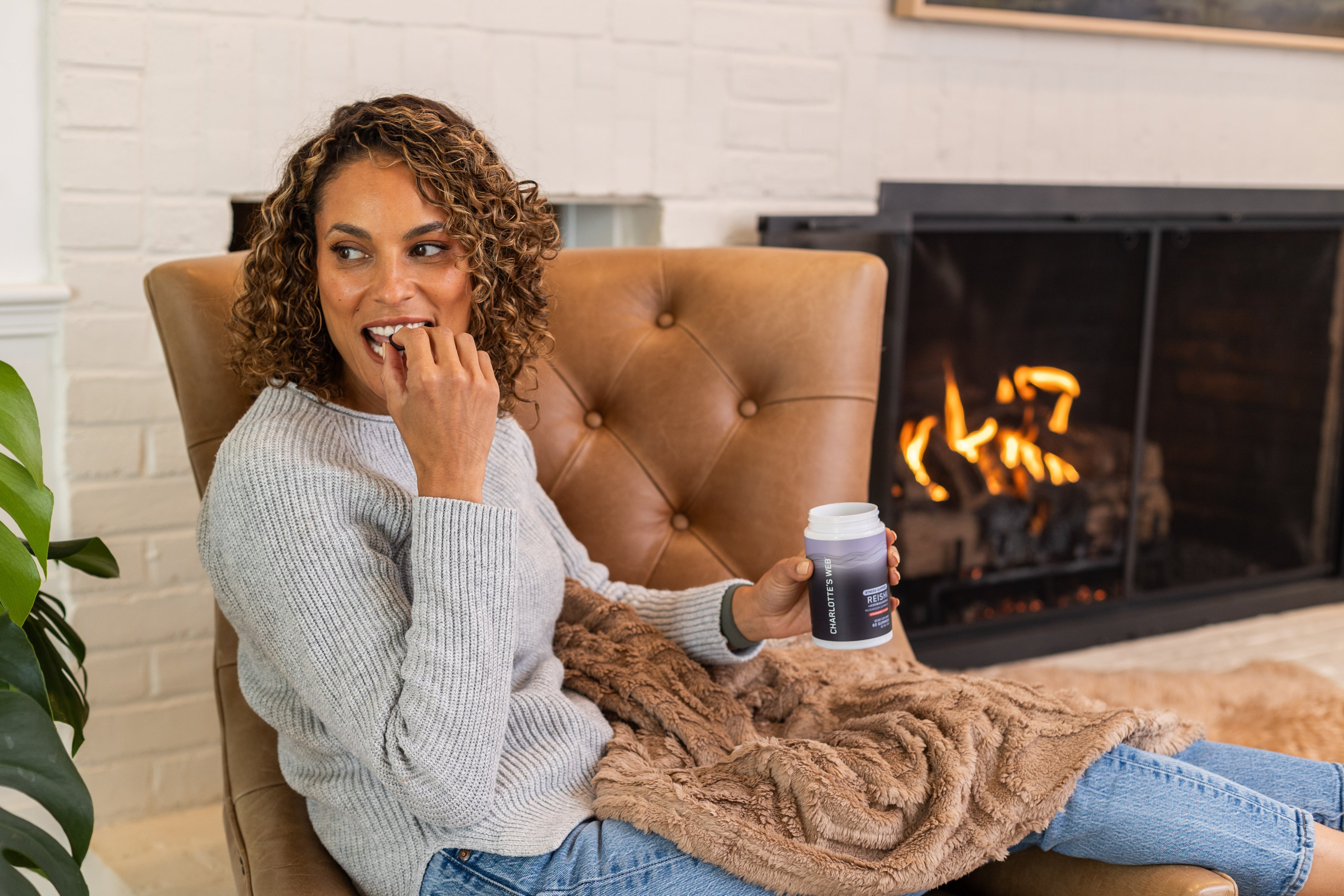 Smiling woman relaxing in a cozy chair by the fireplace, enjoying Charlotte's Web™ Stress Support gummies.